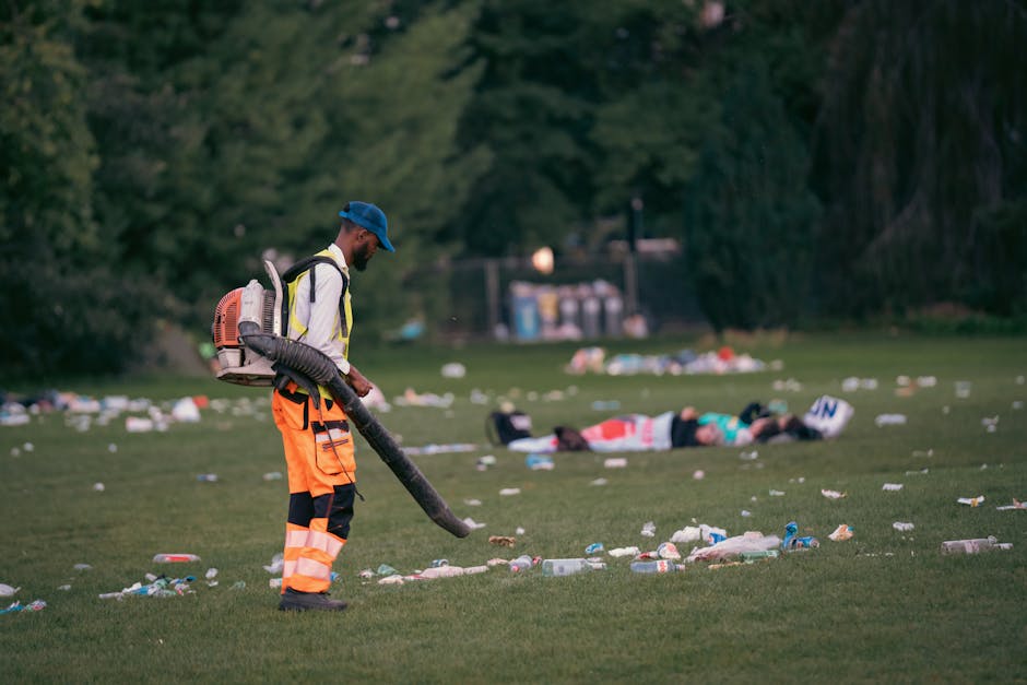 A man wearing a blue cap, high-visibility jacket with reflective stripes, and bright orange trousers is outdoor, operating a leaf blower or vacuum cleaner to clear litter from a grassy park area. The
